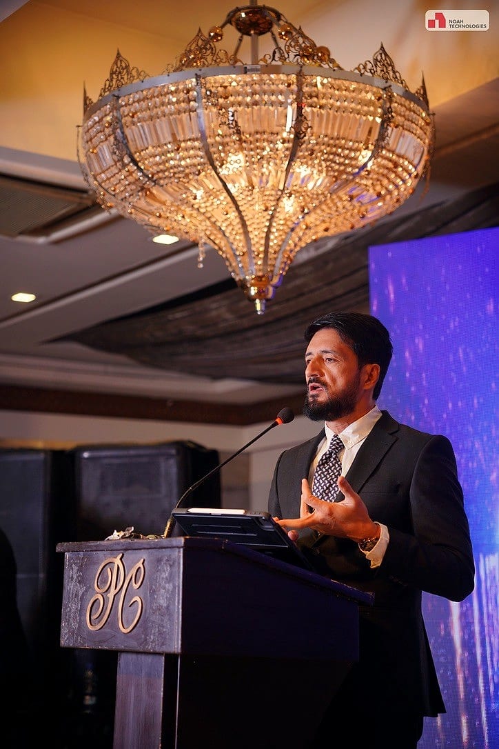 Waqas Khan Pitafi speaking at a podium beneath a chandelier at a technology event